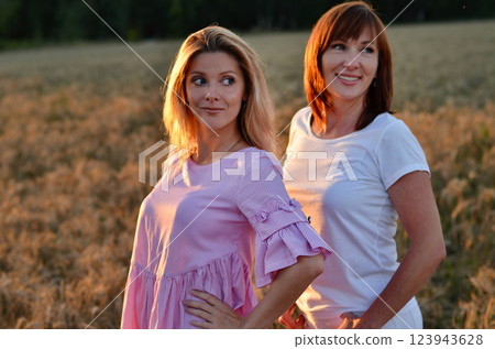 Two women enjoying a joyful moment in a golden wheat field during sunset while wearing bright summer outfits in a rural setting 123943628