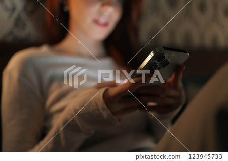 Close-up cropped shot of woman relaxing in dark bedroom, scrolling social media on smartphone, screen warm glow illuminating face. Calm female browsing Internet using mobile phone looking to screen. 123945733