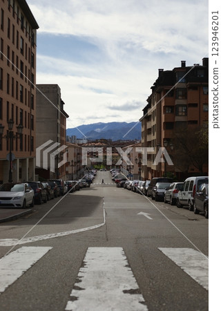 Color photo of an urban street with parked cars, distant mountains, and a single pedestrian crossing the road under a cloudy sky Color photo of an urban street with parked cars, distant mountains, and a single pedestrian crossing the road under a cloudy sky 123946201