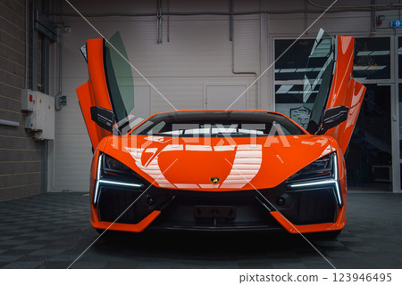 A vibrant orange Lamborghini Revuelto is parked indoors with scissor doors open, showcasing its sleek design against a polished concrete floor and industrial walls. 123946495