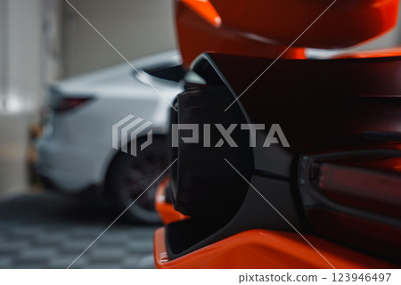 The image captures the rear of a Lamborghini Revuelto, focusing on its orange color, rear light, and exhaust. A blurred white car is in the garage background. 123946497