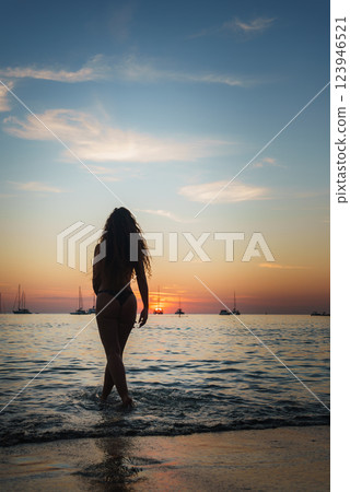 A serene beach scene at sunset in Phuket or Koh Phi Phi, Thailand, shows a woman entering calm waters with silhouetted sailboats on the horizon. A serene beach scene at sunset in Phuket or Koh Phi Phi, Thailand, shows a woman entering calm waters with silhouetted sailboats on the horizon. 123946521