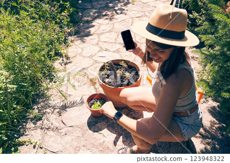 A brunette woman looks at her watch while holding a smartphone with a black screen against the background of a backyard garden. Pot with young plants, work in the summer garden. High quality photo A brunette woman looks at her watch while holding a smartphone with a black screen against the background of a backyard garden. Pot with young plants, work in the summer garden. High quality photo 123948322