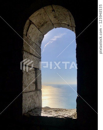 Sea view through the oval window of the stone tower. Sea surface illuminated by the evening sun. 123948535