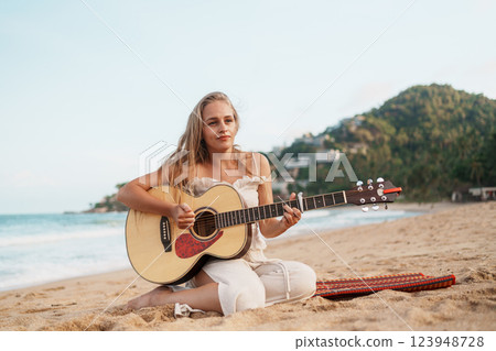 A Woman is Playing a Guitar while Relaxing on the Beach during a Stunning Sunset A Woman is Playing a Guitar while Relaxing on the Beach during a Stunning Sunset 123948728