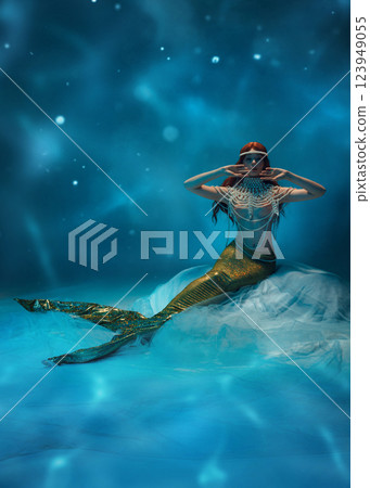 Mystical underwater mermaid, redhaired woman with body covered in pearls, sitting deep in ocean against water-like wavy background. 123949055