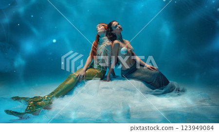 Two young women dressed like mermaids with stylish pearl accessories sitting with eyes closed, executing calmness against blue background 123949084