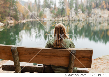 Lonely 30s blonde woman is sitting on the bench of a forest lake. Autumn landscape, calmness, tranquility, travel concept. A woman on the bank of a lake is enjoying the nature. High quality photo. 123949158