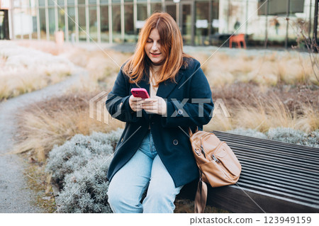 Happy woman sitting on the bench and chatting on phone outdoors. Urban, people concept. Smiling 30s redhead woman text messaging on smart phone at sunny day on the city street, spring time 123949159