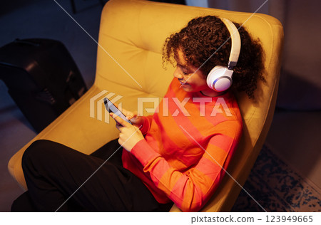 Smiling African woman is sitting in armchair and using phone. Black girl is listening to music and relaxing at home. Top view. Smiling African woman is sitting in armchair and using phone. Black girl is listening to music and relaxing at home. Top view. 123949665