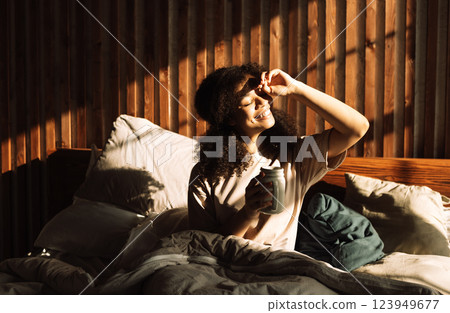 Charming mixed-race teenager is laughing and holding cup of coffee while sitting in bed Charming mixed-race teenager is laughing and holding cup of coffee while sitting in bed 123949677