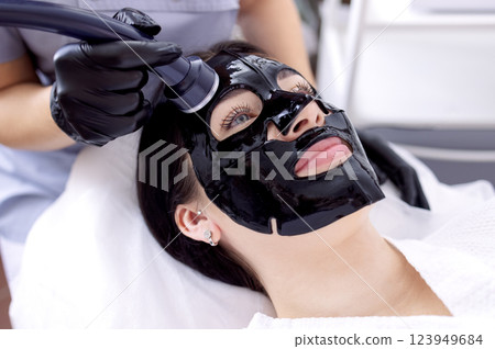 Close-up of a young woman in a black cosmetic mask at a spa procedure. A cosmetologist performs a cosmetic procedure using an ultrasound device. Close-up of a young woman in a black cosmetic mask at a spa procedure. A cosmetologist performs a cosmetic procedure using an ultrasound device. 123949684