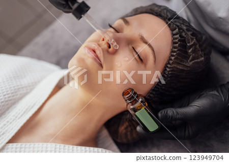Beautician applies serum from pipette in gloves to the face of client in spa salon Beautician applies serum from pipette in gloves to the face of client in spa salon 123949704