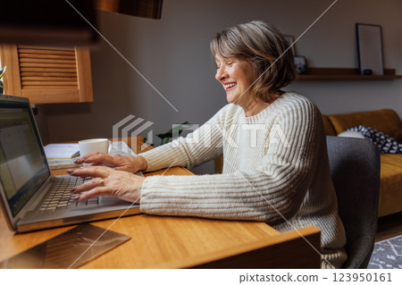 An elderly woman in casual clothes is working on a laptop. The laughing mature lady is typing on a computer. The pensioner smiles and looks at the laptop display. 123950161
