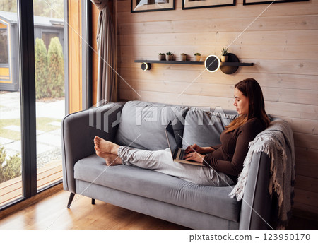A young woman in casual loungewear relaxes on the sofa and working on her laptop. The lady is comfortably sitting with her legs stretched out and making online purchases at home. 123950170