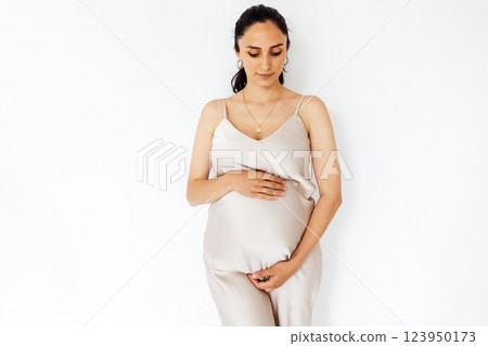 A young pregnant woman stands and gently holds her belly. White isolated background. A charming expectant mother in a white dress. 123950173