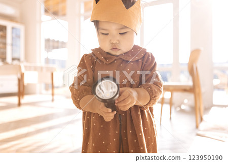 Cute Asian babe is playing with a flashlight at home. A little Korean girl in a dress and with a toy crown on her head in the living room. Cute Asian babe is playing with a flashlight at home. A little Korean girl in a dress and with a toy crown on her head in the living room. 123950190