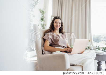 A calm young freelance woman in casual clothes is sitting in an armchair with a notebook and laptop in the bedroom A calm young freelance woman in casual clothes is sitting in an armchair with a notebook and laptop in the bedroom 123950193