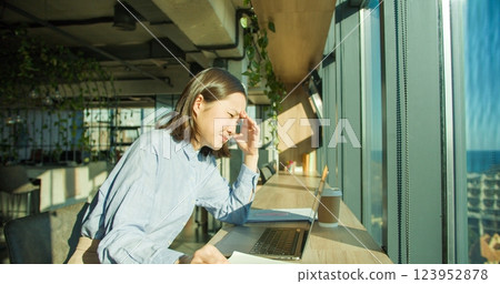 A thoughtful young woman in a modern office struggles with fatigue as sunlight streams through large windows, surrounded by a laptop, coffee, and notepad 123952878