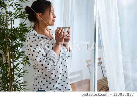 Woman Enjoying Beverage in Cozy Home While Contemplating at the Window 123953853