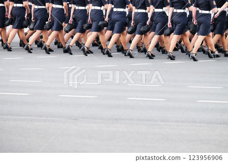 A group of women in blue uniforms is marching in synchronized formation on an urban street. They wear high heels and are participating in a public ceremony with an organized display. 123956906