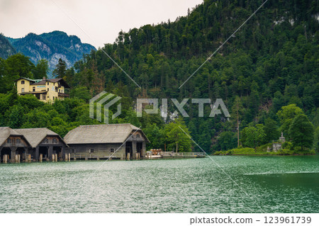 Vintage wooden boathouses line the calm, green waters, surrounded by dense trees and towering mountains under a cloudy sky, creating a tranquil atmosphere in nature. Konigssee lake Bavaria German Alps Vintage wooden boathouses line the calm, green waters, surrounded by dense trees and towering mountains under a cloudy sky, creating a tranquil atmosphere in nature. Konigssee lake Bavaria German Alps 123961739