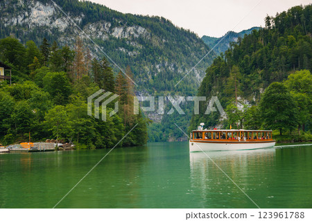 A boat gracefully moves through the calm waters of a beautiful lake, bordered by dramatic green hills and dense forests under a cloudy sky. The peaceful setting invites relaxation. Konigssee Bavaria A boat gracefully moves through the calm waters of a beautiful lake, bordered by dramatic green hills and dense forests under a cloudy sky. The peaceful setting invites relaxation. Konigssee Bavaria 123961788