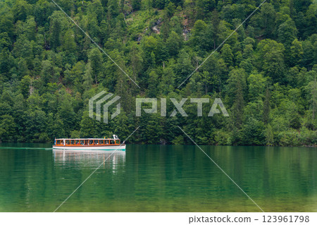 A boat gracefully moves through the calm waters of a beautiful lake, bordered by dramatic green hills and dense forests under a cloudy sky. The peaceful setting invites relaxation. Konigssee Bavaria 123961798