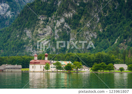 A white church with red domes stands by the tranquil waters of a lake, surrounded by lush greenery and majestic mountains on a sunny day. Konigssee lake Bavaria German Alps Berchtesgaden church by the A white church with red domes stands by the tranquil waters of a lake, surrounded by lush greenery and majestic mountains on a sunny day. Konigssee lake Bavaria German Alps Berchtesgaden church by the 123961807