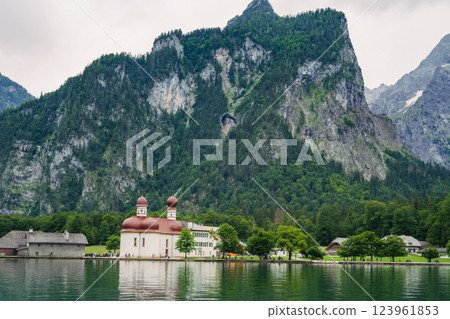 A white church with red domes stands by the tranquil waters of a lake, surrounded by lush greenery and majestic mountains on a sunny day. Konigssee lake Bavaria German Alps Berchtesgaden church by the A white church with red domes stands by the tranquil waters of a lake, surrounded by lush greenery and majestic mountains on a sunny day. Konigssee lake Bavaria German Alps Berchtesgaden church by the 123961853