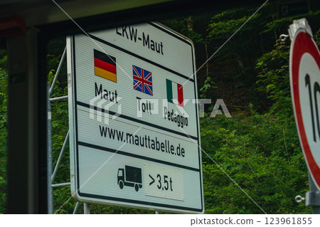 A toll sign is prominently displayed beside a highway in Germany, featuring information in German, English, and Italian. The area is lush and green, indicating a beautiful landscape. A toll sign is prominently displayed beside a highway in Germany, featuring information in German, English, and Italian. The area is lush and green, indicating a beautiful landscape. 123961855