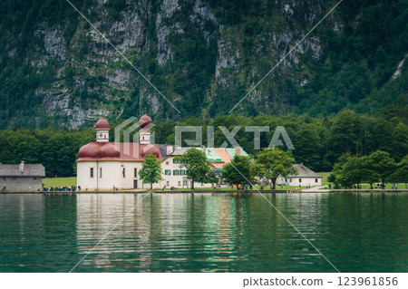 A white church with red domes stands by the tranquil waters of a lake, surrounded by lush greenery and majestic mountains on a sunny day. Konigssee lake Bavaria German Alps Berchtesgaden church by the 123961856