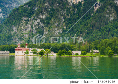 A white church with red domes stands by the tranquil waters of a lake, surrounded by lush greenery and majestic mountains on a sunny day. Konigssee lake Bavaria German Alps Berchtesgaden church by the 123961916