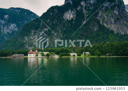 A white church with red domes stands by the tranquil waters of a lake, surrounded by lush greenery and majestic mountains on a sunny day. Konigssee lake Bavaria German Alps Berchtesgaden church by the A white church with red domes stands by the tranquil waters of a lake, surrounded by lush greenery and majestic mountains on a sunny day. Konigssee lake Bavaria German Alps Berchtesgaden church by the 123961953