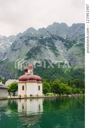 A white church with red domes stands by the tranquil waters of a lake, surrounded by lush greenery and majestic mountains on a sunny day. Konigssee lake Bavaria German Alps Berchtesgaden church by the A white church with red domes stands by the tranquil waters of a lake, surrounded by lush greenery and majestic mountains on a sunny day. Konigssee lake Bavaria German Alps Berchtesgaden church by the 123961997