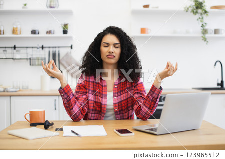 Multiracial woman meditating at home kitchen table with computer, feeling calm and relaxed. Focus on peace and balance, practicing mindfulness for stress management, casual indoor lifestyle concept. 123965512