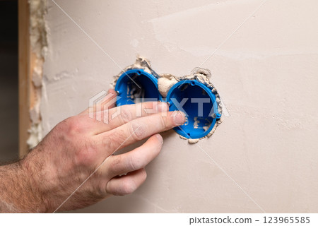 Electrician installing blue socket boxes into a plastered wall during renovation or construction. 123965585