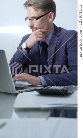Businessman wearing blue suit is thinking while working with laptop, calculator and documents at the glass table in office set. Low lighting, vertical portrait. Business people and audit concept 123971118
