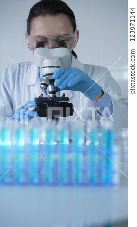 Female scientist wearing lab coat and protective blue gloves using microscope doing research in modern laboratory, portrait view. Low key lighting photo. Science and Medicine Female scientist wearing lab coat and protective blue gloves using microscope doing research in modern laboratory, portrait view. Low key lighting photo. Science and Medicine 123971144