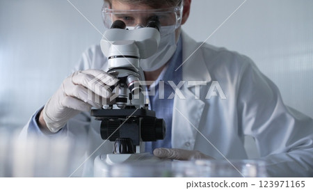 Man scientist researcher wearing white gloves, protective glasses and medical mask is using microscope in modern laboratory, portrait view. Low key lighting photo. Science and Medicine Man scientist researcher wearing white gloves, protective glasses and medical mask is using microscope in modern laboratory, portrait view. Low key lighting photo. Science and Medicine 123971165