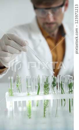 Man scientist wearing a lab coat, white gloves and protective glasses, is dripping liquid from a pipette into a test tube with a green plant inside, vertical portrait. Science and medicine concepts 123971213