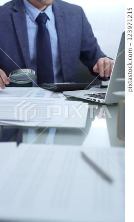 Businessman wearing blue suit is analyzing financial reports with magnifying glass and using calculator in a professional office setting. Low lighting, close-up vertical view. Business people, audit 123971215