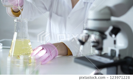 A scientist, wearing a lab coat and pink protective gloves, is pouring a yellow oily liquid from one beaker to another near microscope in laboratory, close up. Medicine and science A scientist, wearing a lab coat and pink protective gloves, is pouring a yellow oily liquid from one beaker to another near microscope in laboratory, close up. Medicine and science 123971228