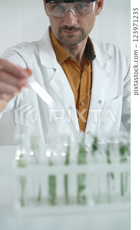 A male science researcher, wearing a lab coat and protective glasses, drips liquid from a pipette into a test tube containing a green plant. His face is focused, captured in a vertical portrait 123971235