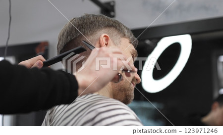 Professional barber trimming blond heard male client's hair with scissors and comb, creating precise modern hairstyle inside contemporary grooming salon Professional barber trimming blond heard male client's hair with scissors and comb, creating precise modern hairstyle inside contemporary grooming salon 123971246
