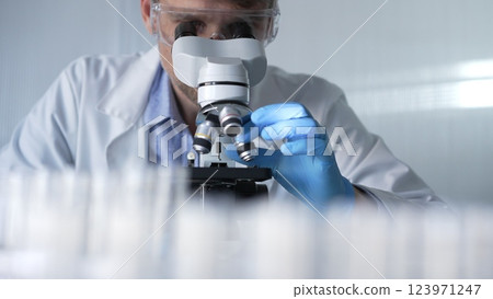 Male scientist researcher wearing blue gloves, protective glasses is using microscope in modern laboratory, portrait view. Low key lighting photo. Science and Medicine concepts Male scientist researcher wearing blue gloves, protective glasses is using microscope in modern laboratory, portrait view. Low key lighting photo. Science and Medicine concepts 123971247