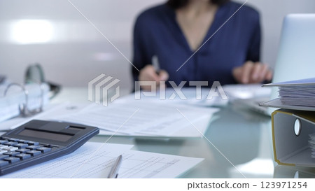 Black calculator, pen and documents are lying on accountant's desk. Businesswoman wearing blue dress is working with documents on the background. Business and audit concept Black calculator, pen and documents are lying on accountant's desk. Businesswoman wearing blue dress is working with documents on the background. Business and audit concept 123971254