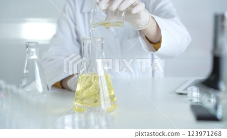 A scientist, wearing a lab coat and white protective gloves, is pouring a yellow oily liquid from one beaker to another in laboratory, close up. Medicine and science concept 123971268
