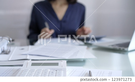White calculator, pen and documents are lying on accountant's desk. Businesswoman wearing blue dress is working with documents on the background. Business and audit concept 123971273