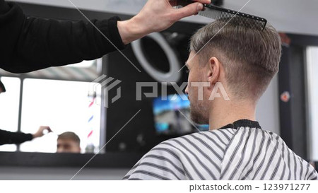 Barber carefully combing hair, client wearing protective cape, preparing for precision styling inside sleek contemporary barbershop setting Barber carefully combing hair, client wearing protective cape, preparing for precision styling inside sleek contemporary barbershop setting 123971277